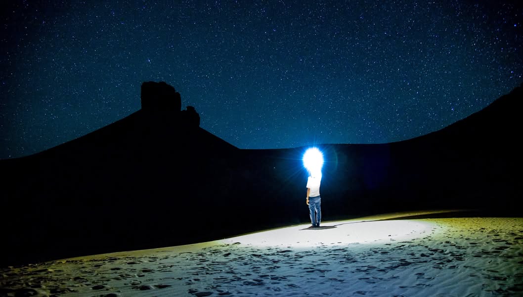A person shines a light beneath a star-filled sky, between sand dunes and silhouetted rocks.