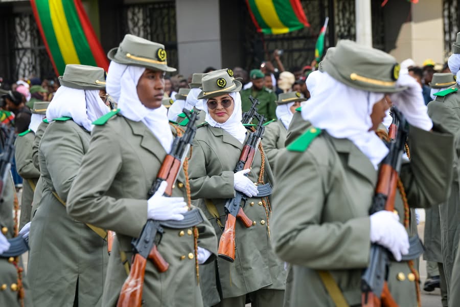 Women in ceremonial military uniforms march past Mauritanian flags during a parade.