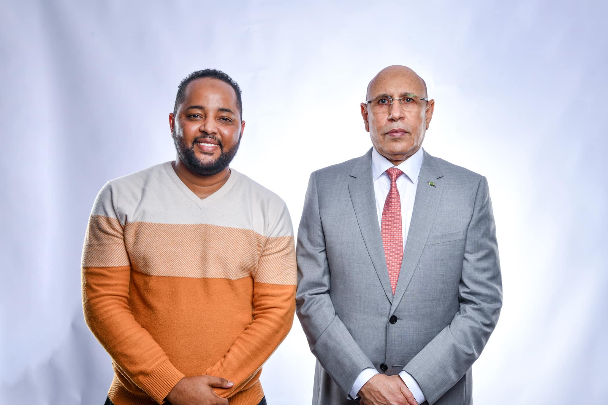 Two men pose against a pale background, one in a striped sweater and the other in a grey suit.
